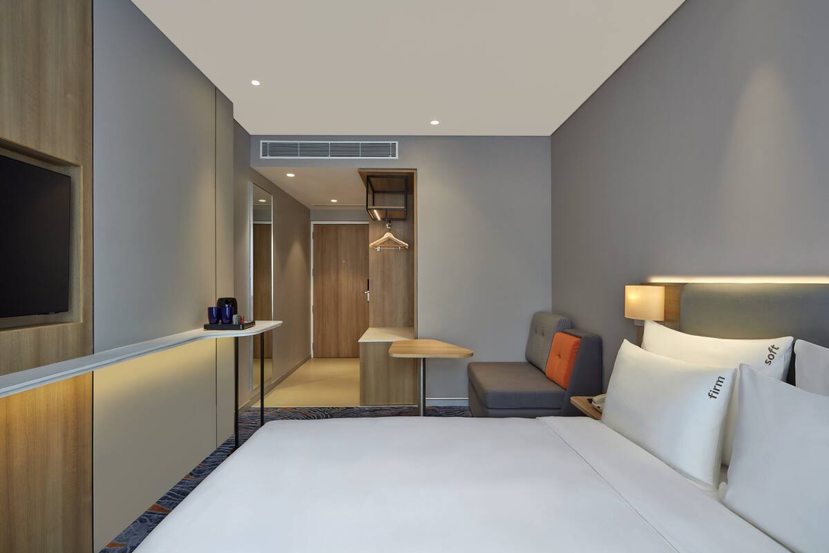 A modern hotel room with a large bed, a desk, a seating area, and a TV mounted on the wall.