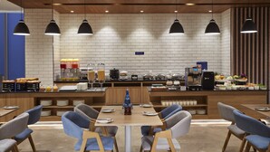 Een modern café met een bakstenen muur, houten plafond en zwarte hanglampen. Er zijn tafels en stoelen, plus een balie met eten en drinken.