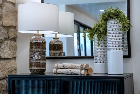 Un intérieur moderne avec un mur en pierre, un meuble bleu foncé, deux vases à texture, un miroir et un vase blanc orné de verdure.