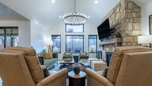 A spacious living room with leather armchairs, a stone fireplace, and large windows offering a view of trees. - Redtail Rendezvous with Hot Tub (Driggs)