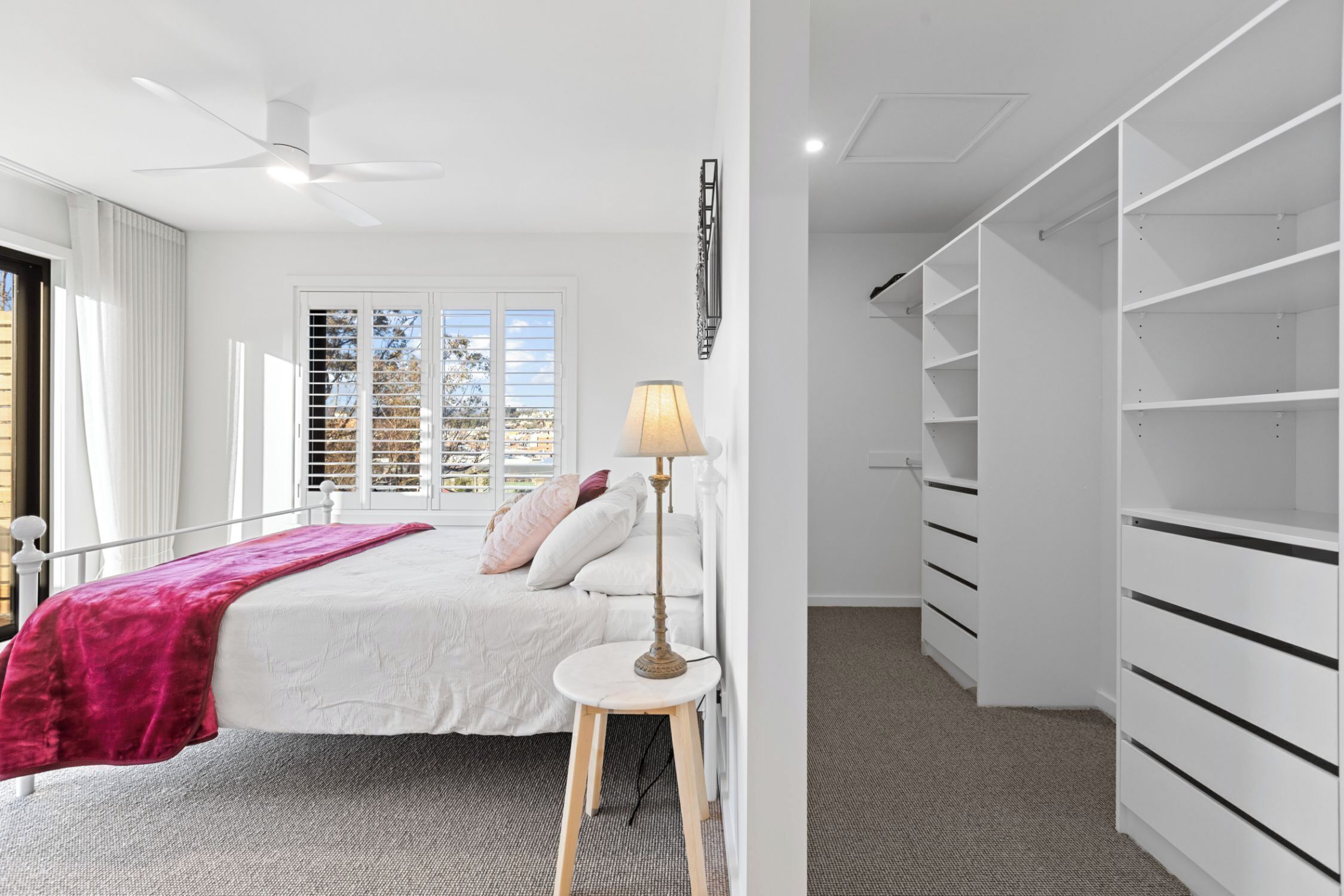 A bedroom with a bed, a bedside table with a lamp, a closet with shelves, and a window with blinds.