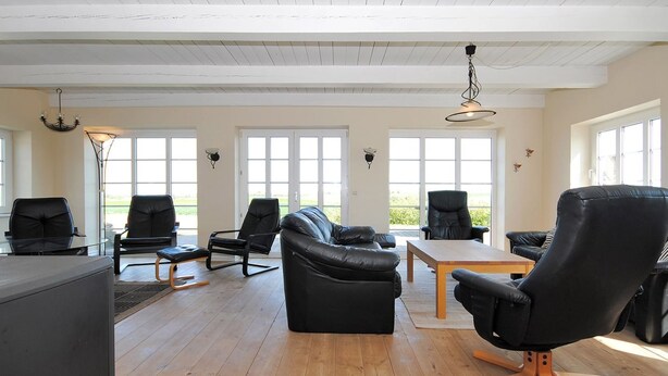 Un salon spacieux avec un canapé en cuir noir, une table basse en bois et des fauteuils en cuir noir. De grandes fenêtres laissent entrer la lumière naturelle et offrent une vue sur l’extérieur.
