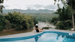 Eine Person sitzt am Pool, im Hintergrund ein Fluss und Berge.