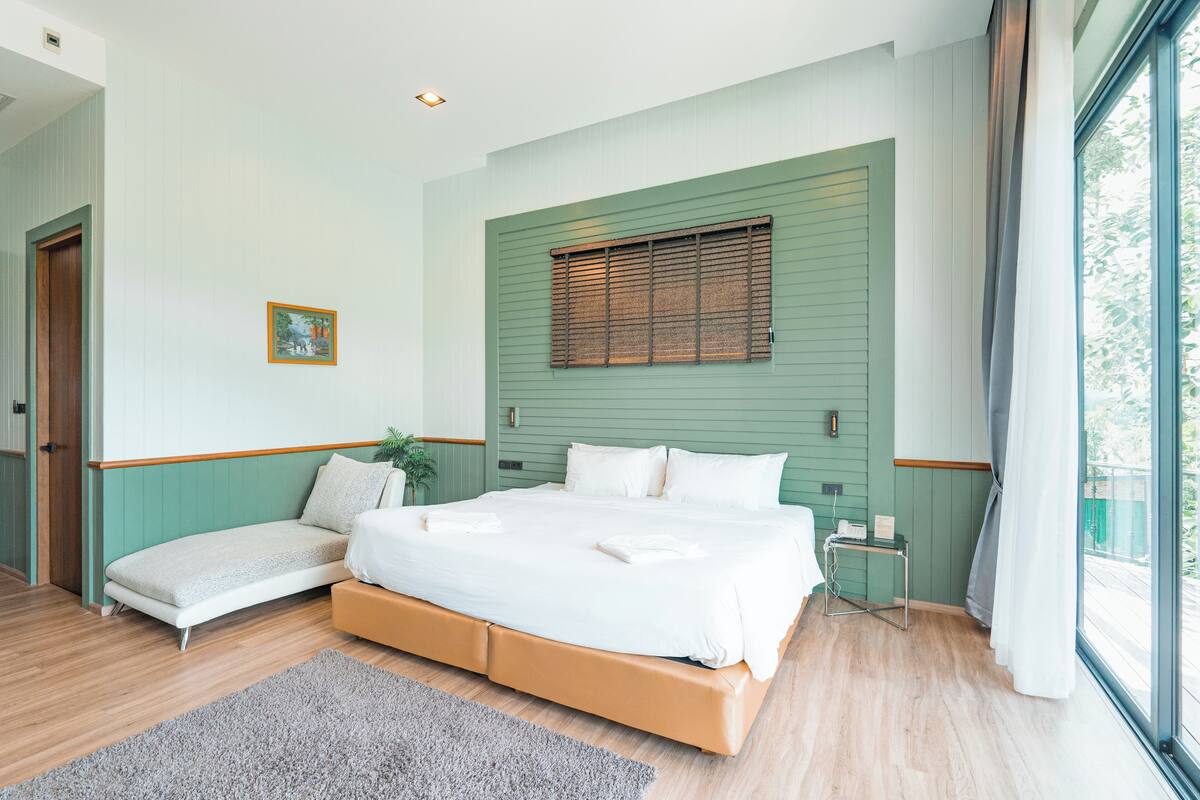 Een slaapkamer met een groot bed, een nachtkastje, een kleine bedlamp en een raam met zonwering, en een deur.