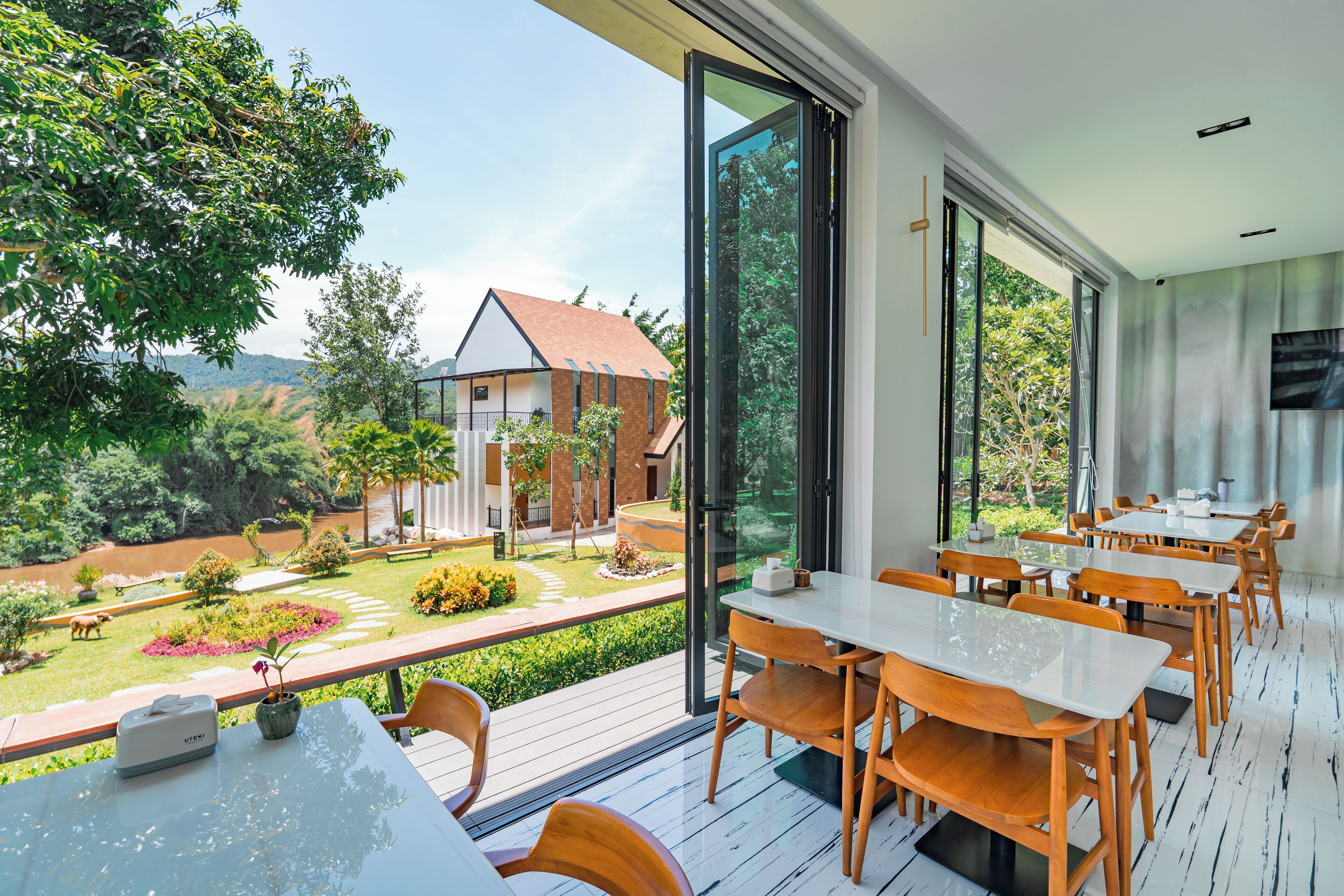 A modern outdoor dining area with large windows overlooking a garden and a house.