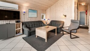Un salon chaleureux avec un canapé, un fauteuil et une table basse. Un téléviseur à écran plat est fixé au mur, ainsi qu’un petit meuble et une cheminée.