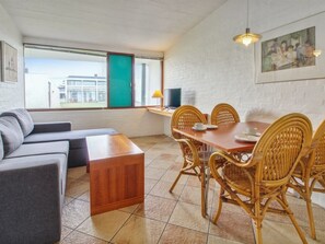 Een moderne woonkamer met een bank, een houten salontafel, een eettafel met rieten stoelen, een televisie en een raam met uitzicht op een gebouw.