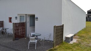 Een modern gebouw met een platte gevel, een deur en een klein buitenterrein met metalen stoelen en een tafel.