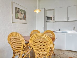 Een eetruimte met rieten stoelen, een houten tafel, een witte baksteenmuur, een ingelijst beeld en een keuken met witte kasten en apparatuur.