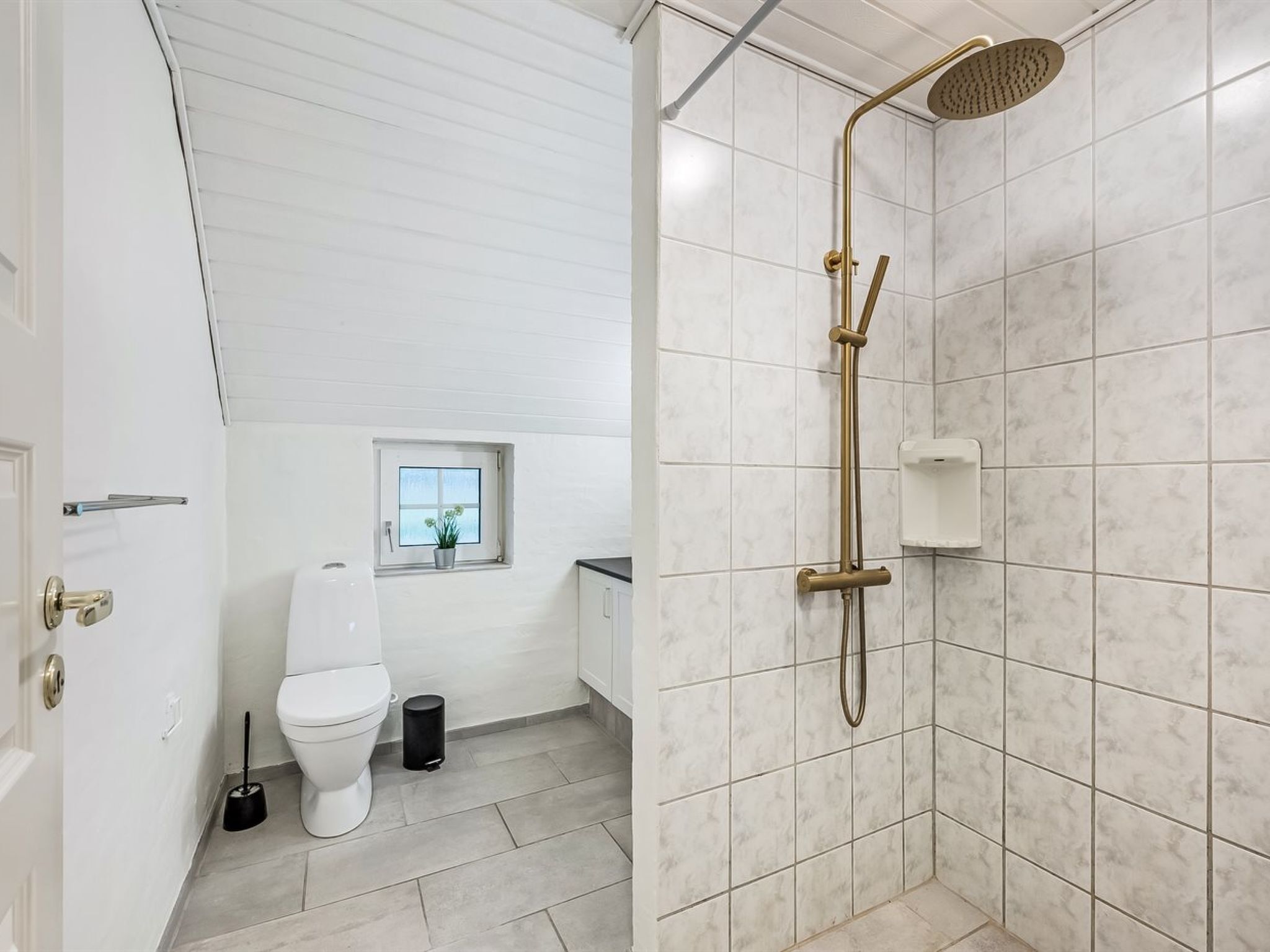 Ein modernes Badezimmer mit Toilette in Weiß, einem kleinen Fenster mit Pflanze und einer Dusche mit goldfarbener Brause.