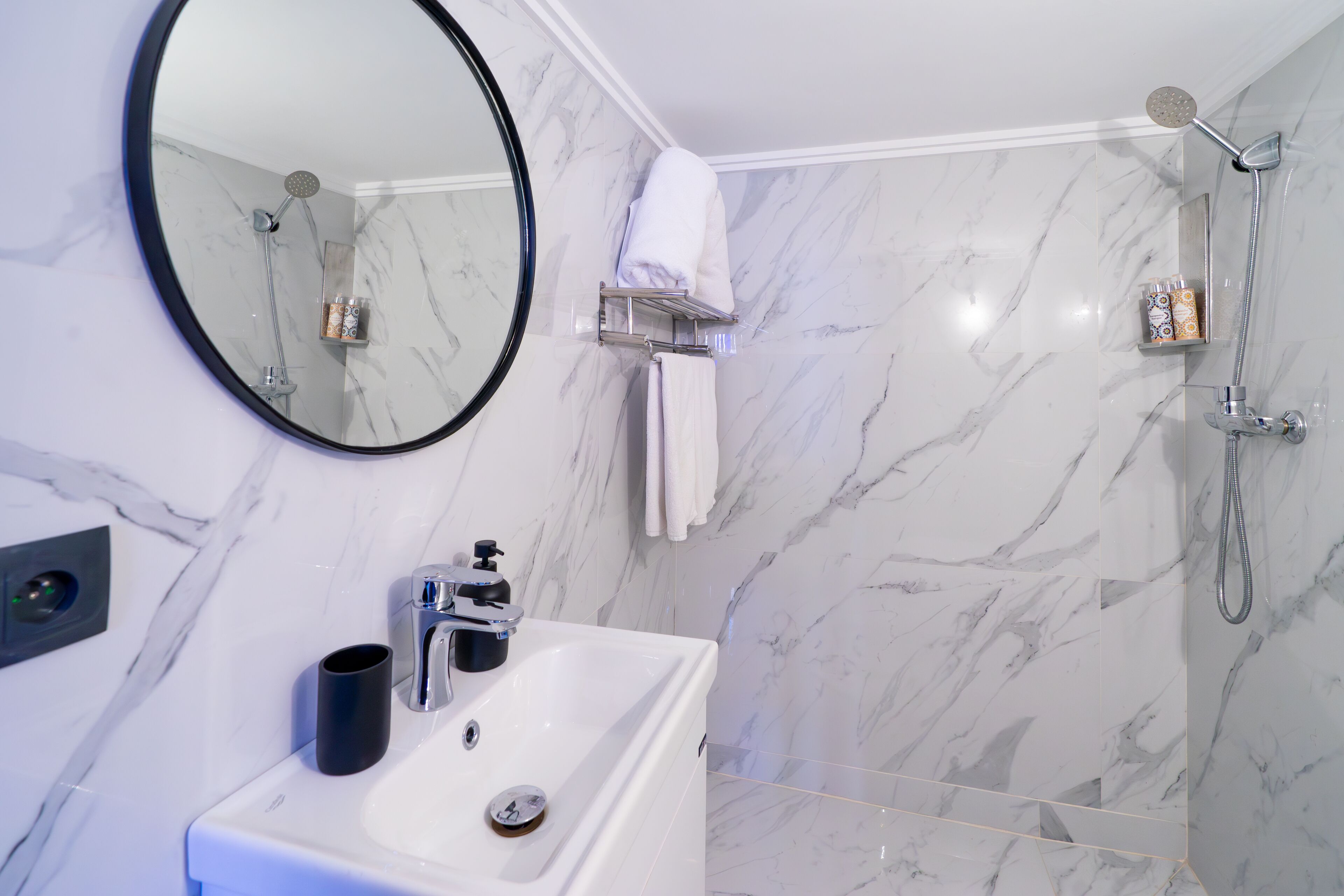 A bathroom with a white sink, a round mirror, a shower with a handheld showerhead, and a towel rack.