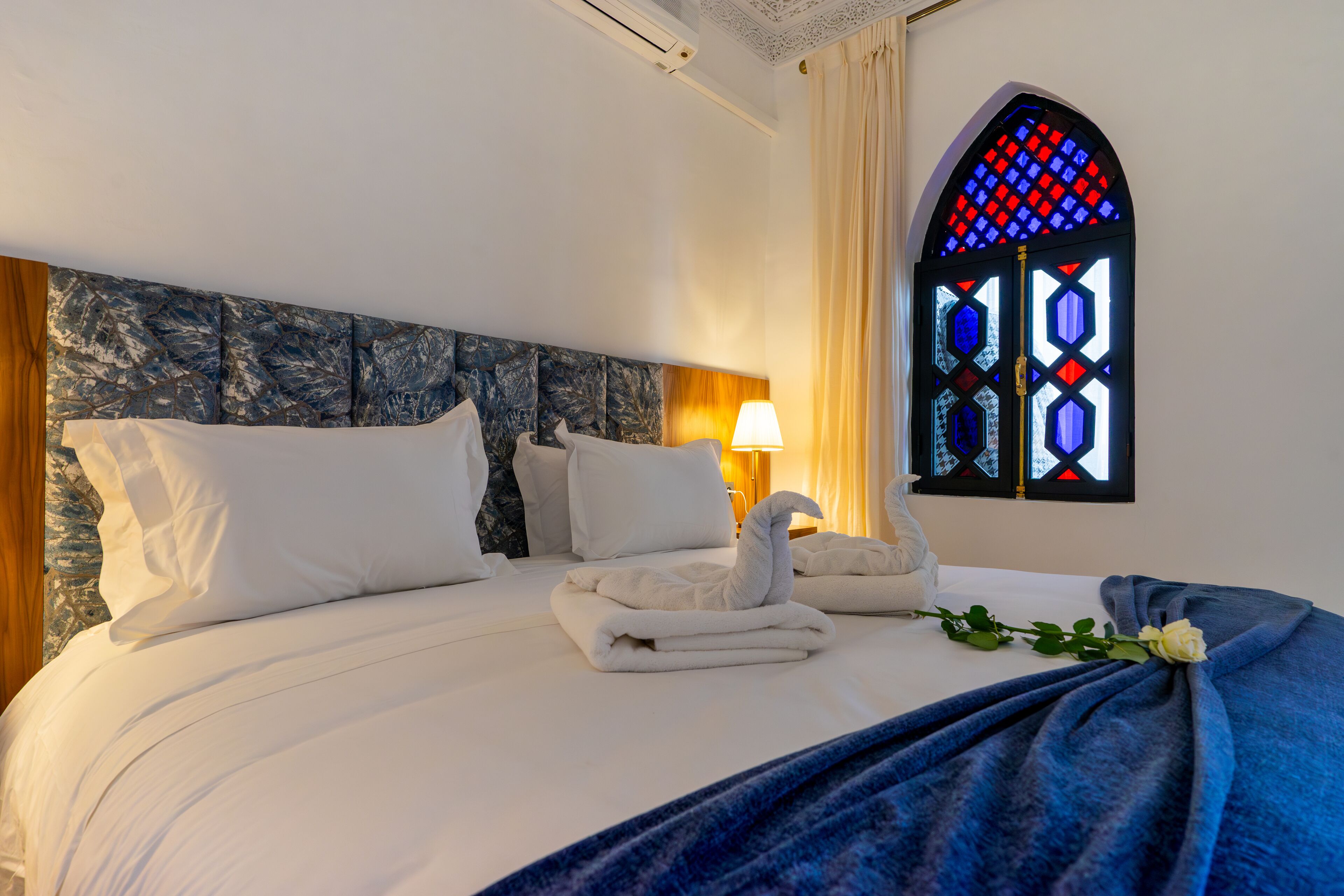 A neatly made bed with white linens and a blue blanket, a wall-mounted lamp, and a stained glass window with a geometric design.