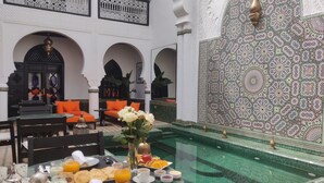 A luxurious indoor pool area with a dining setup featuring a table with breakfast items, surrounded by comfortable seating and decorative elements. - The Grand Riad & Spa  (Marrakech)