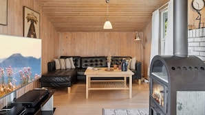 A cozy living room with a wooden ceiling, a black leather sofa, a wooden coffee table, and a wood-burning stove.