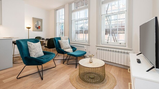A modern living room with two teal armchairs, a round coffee table, a flat-screen TV, and large windows. - Leicester Square  flats by Joivy (London)