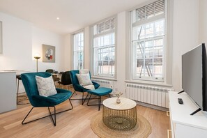 A modern living room with two teal armchairs, a round coffee table, a flat-screen TV, and large windows. - Leicester Square  flats by Joivy (London)