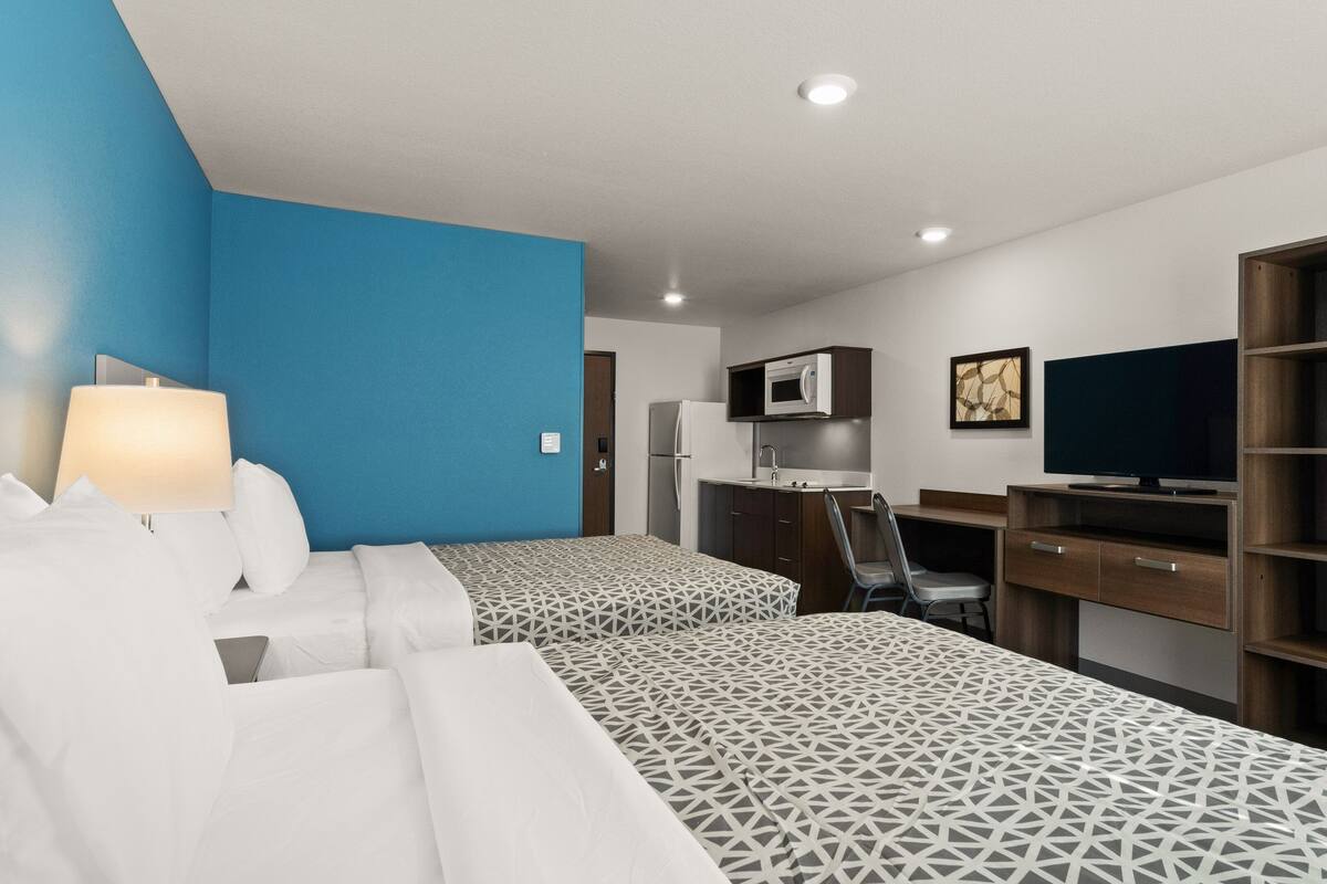 A hotel room with two beds, a desk, a TV, and a kitchenette.