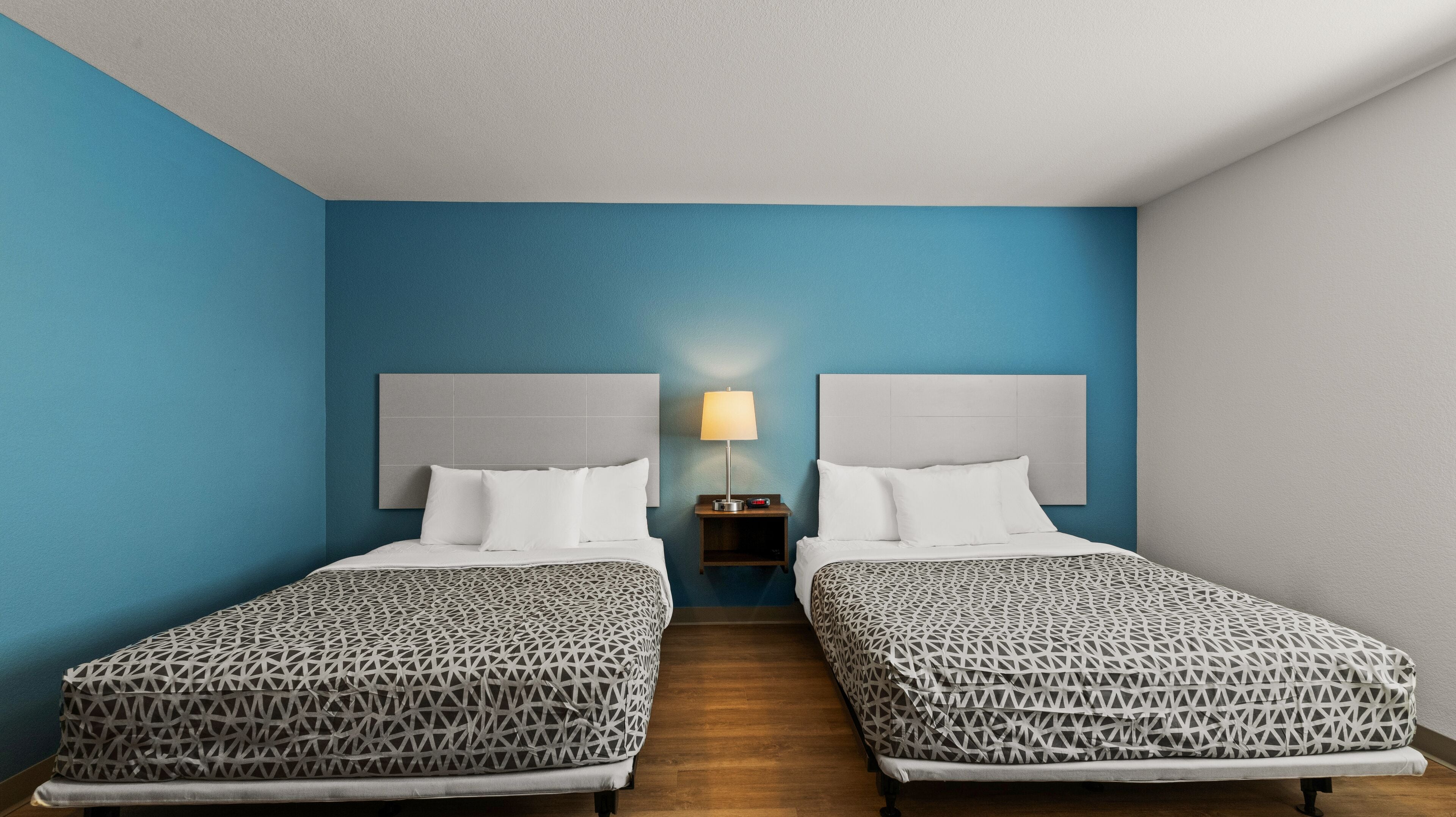 Habitación de hotel con dos camas, una mesita de noche con lámpara y una pared azul decorativa.