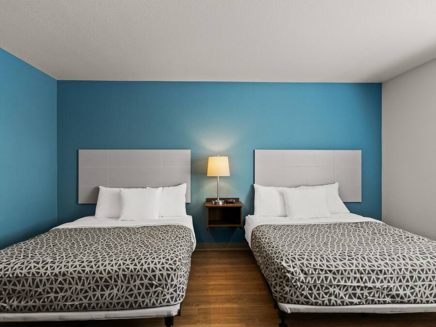 Habitación de hotel con dos camas, una mesita de noche con lámpara y una pared azul decorativa.