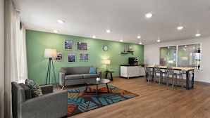 A modern living room with a green accent wall, a grey sofa, a coffee table, and a dining area with chairs and a table. - WoodSpring Suites Greeley (Greeley)