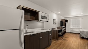 A compact kitchenette with a microwave, sink, and cabinet, a small desk with a chair, and a bed with a patterned cover. - WoodSpring Suites Greeley (Greeley)