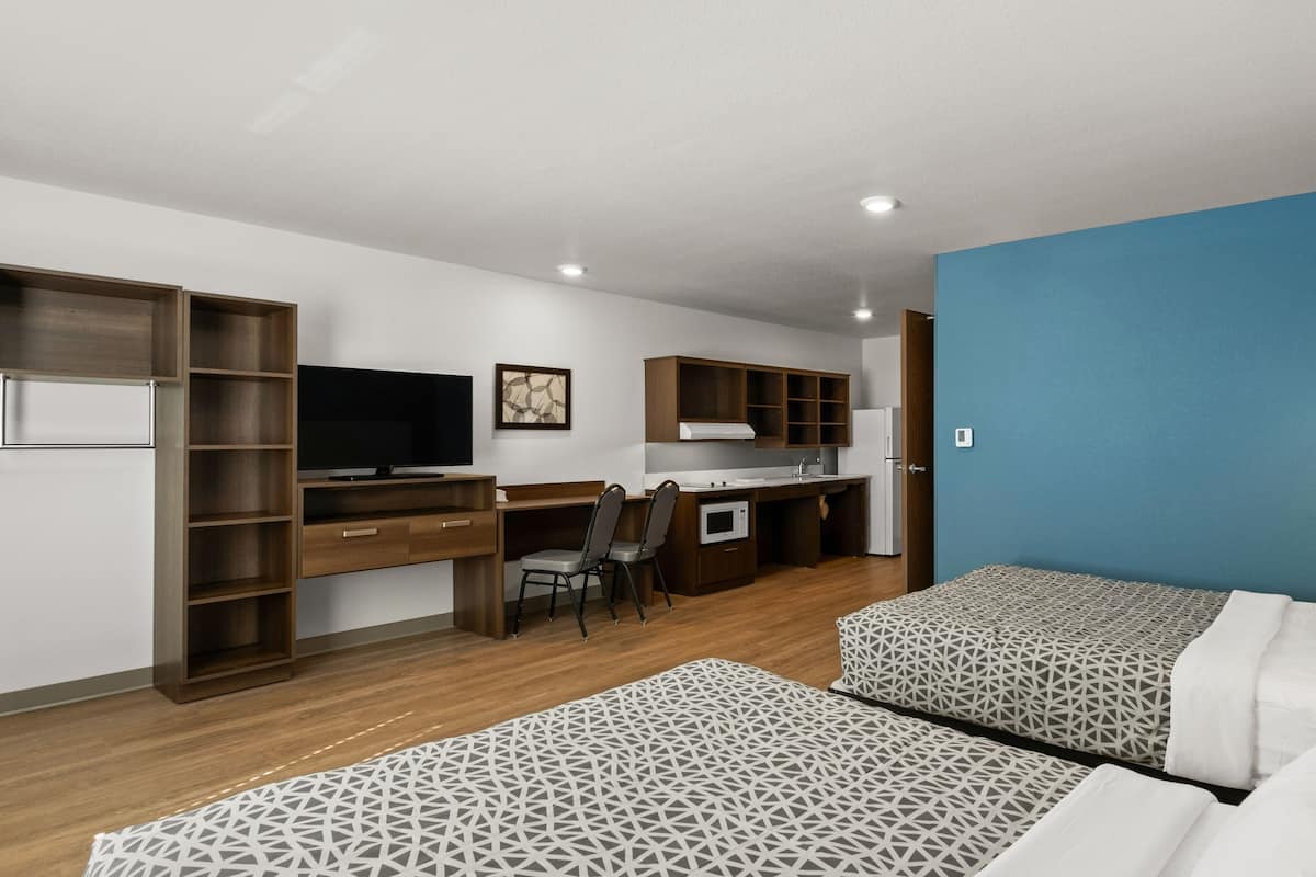 A compact hotel room with two beds, a desk with a chair, a television, a microwave, and a kitchenette.