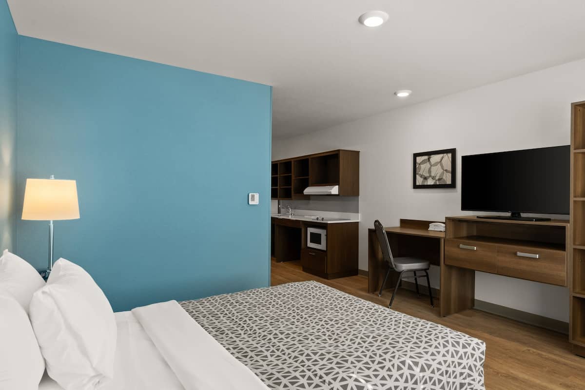 A compact hotel room with a bed, a desk, a kitchenette, and a TV.