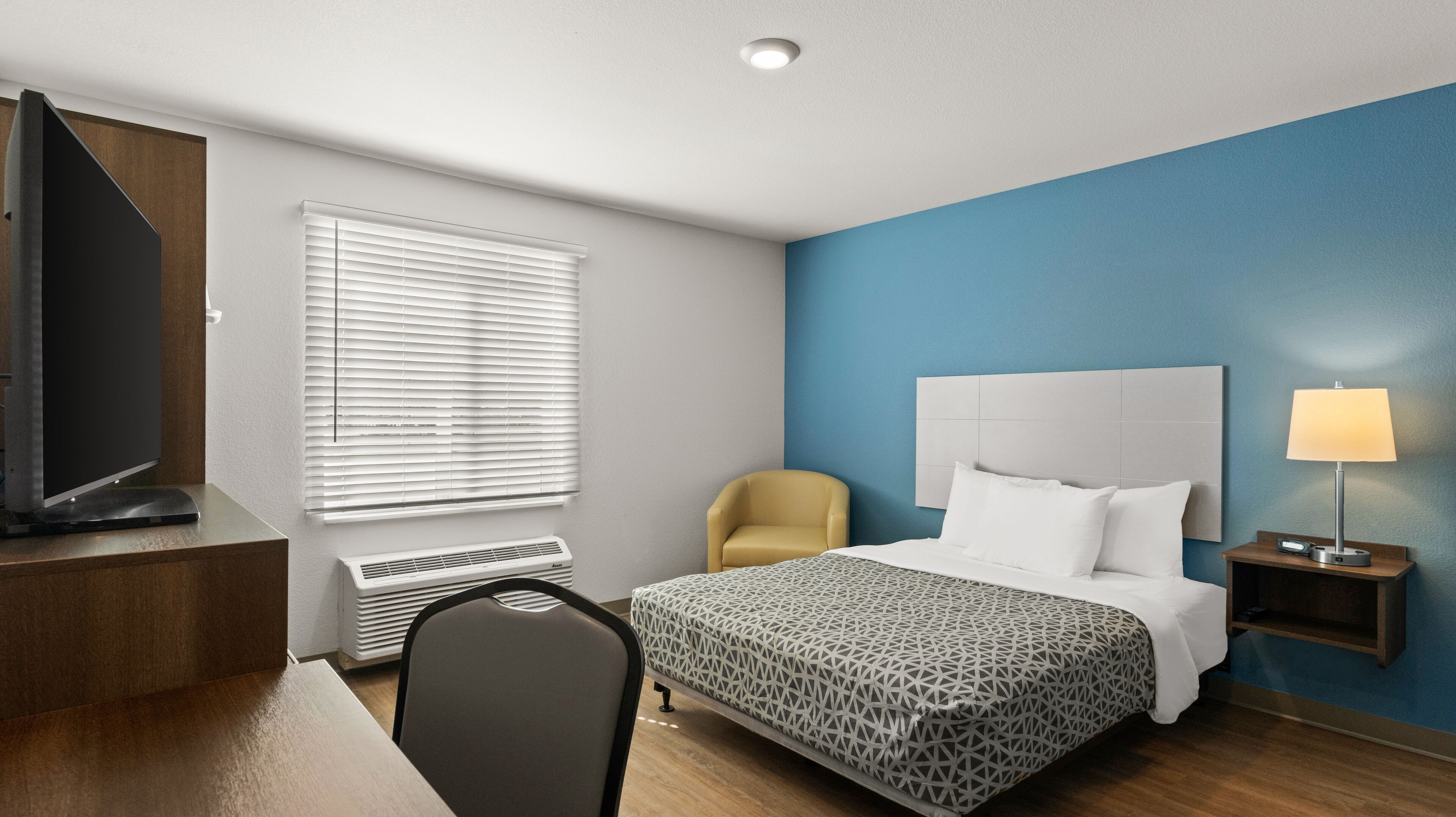A hotel room with a bed, a chair, a desk with a television, a window with blinds, a nightstand with a lamp, and a wall unit with a flat-screen TV.