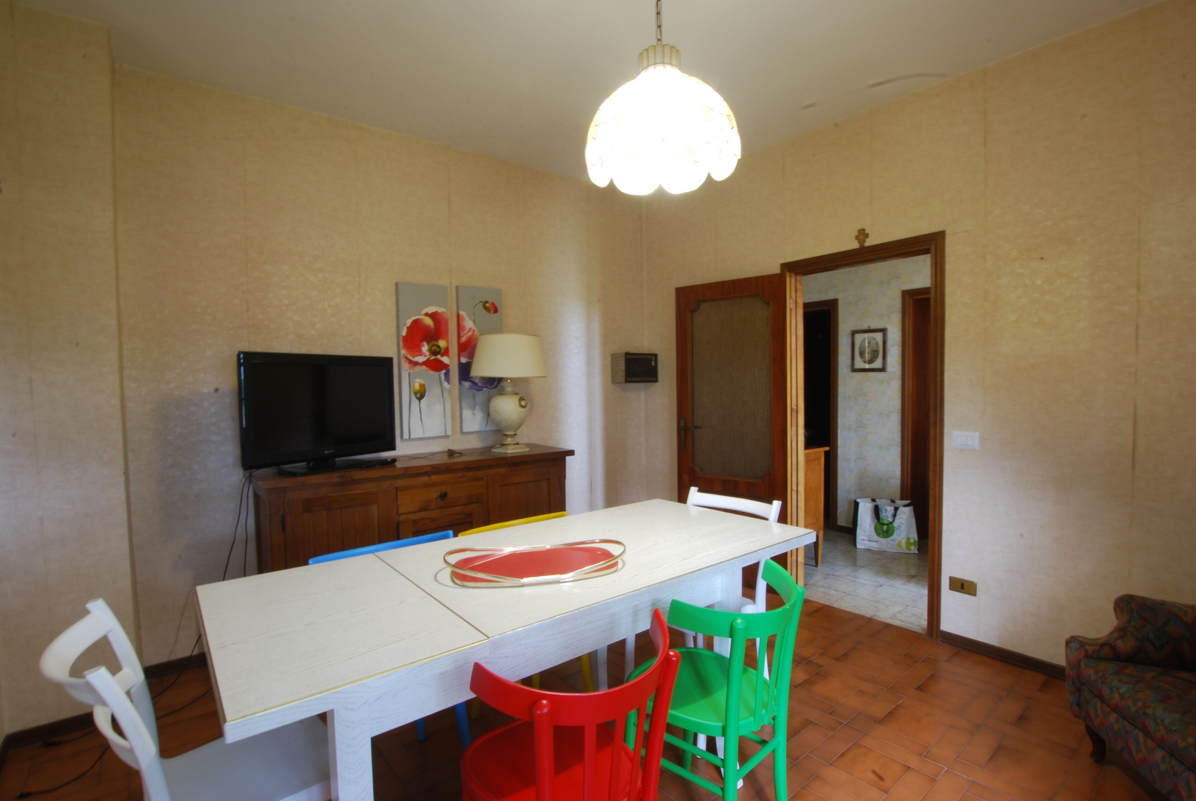 Een eetgedeelte met een witte tafel, rode en groene stoelen, een televisie op een houten kast en een hangende lamp.