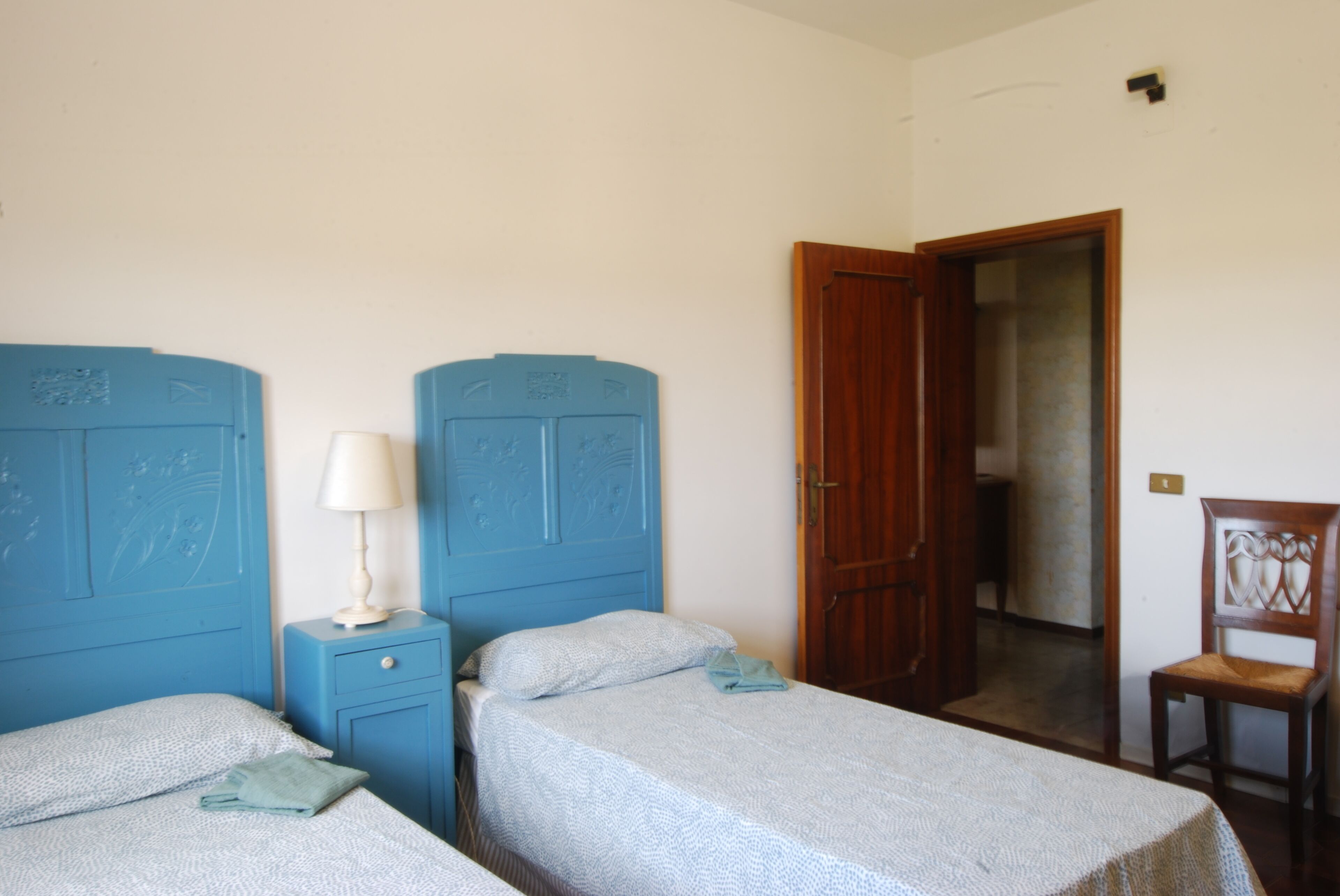 A room with two single beds, a wooden wardrobe, a small bedside table with a lamp, and a wooden chair.