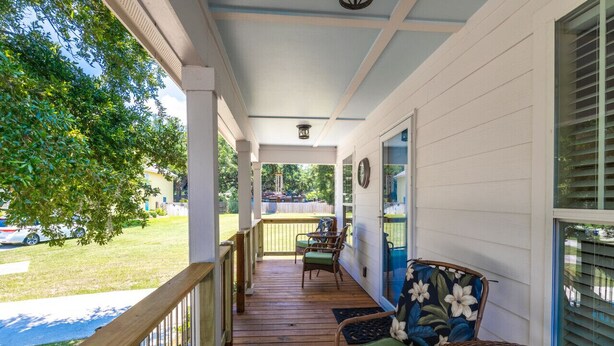 A covered porch with wooden flooring, wicker furniture, and a view of a green lawn and parked cars. - Tranquil Tides Beach Cottage — Walk to the Beach + Relax in Style (Gulfport)