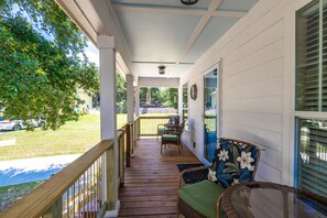 A covered porch with wooden flooring, wicker furniture, and a view of a green lawn and parked cars. - Tranquil Tides Beach Cottage — Walk to the Beach + Relax in Style (Gulfport)