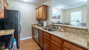 A kitchen with wooden cabinets, a black refrigerator, a granite countertop, a sink, and a toaster. - Tranquil Tides Beach Cottage — Walk to the Beach + Relax in Style (Gulfport)