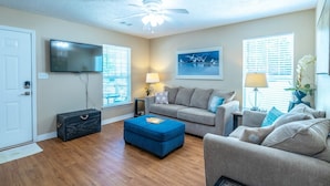 A living room with a sofa, ottoman, ceiling fan, TV, and windows with blinds. - Tranquil Tides Beach Cottage — Walk to the Beach + Relax in Style (Gulfport)