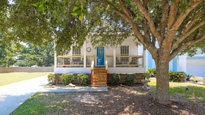 A two-story house with a covered porch, a wooden deck, and a large tree in front. - Tranquil Tides Beach Cottage — Walk to the Beach + Relax in Style (Gulfport)