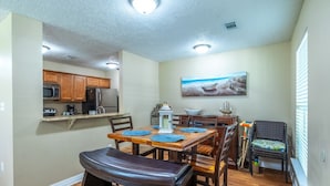 A dining area with a wooden table, chairs, and a bench. There is a kitchen area with cabinets and appliances in the background. - Tranquil Tides Beach Cottage — Walk to the Beach + Relax in Style (Gulfport)