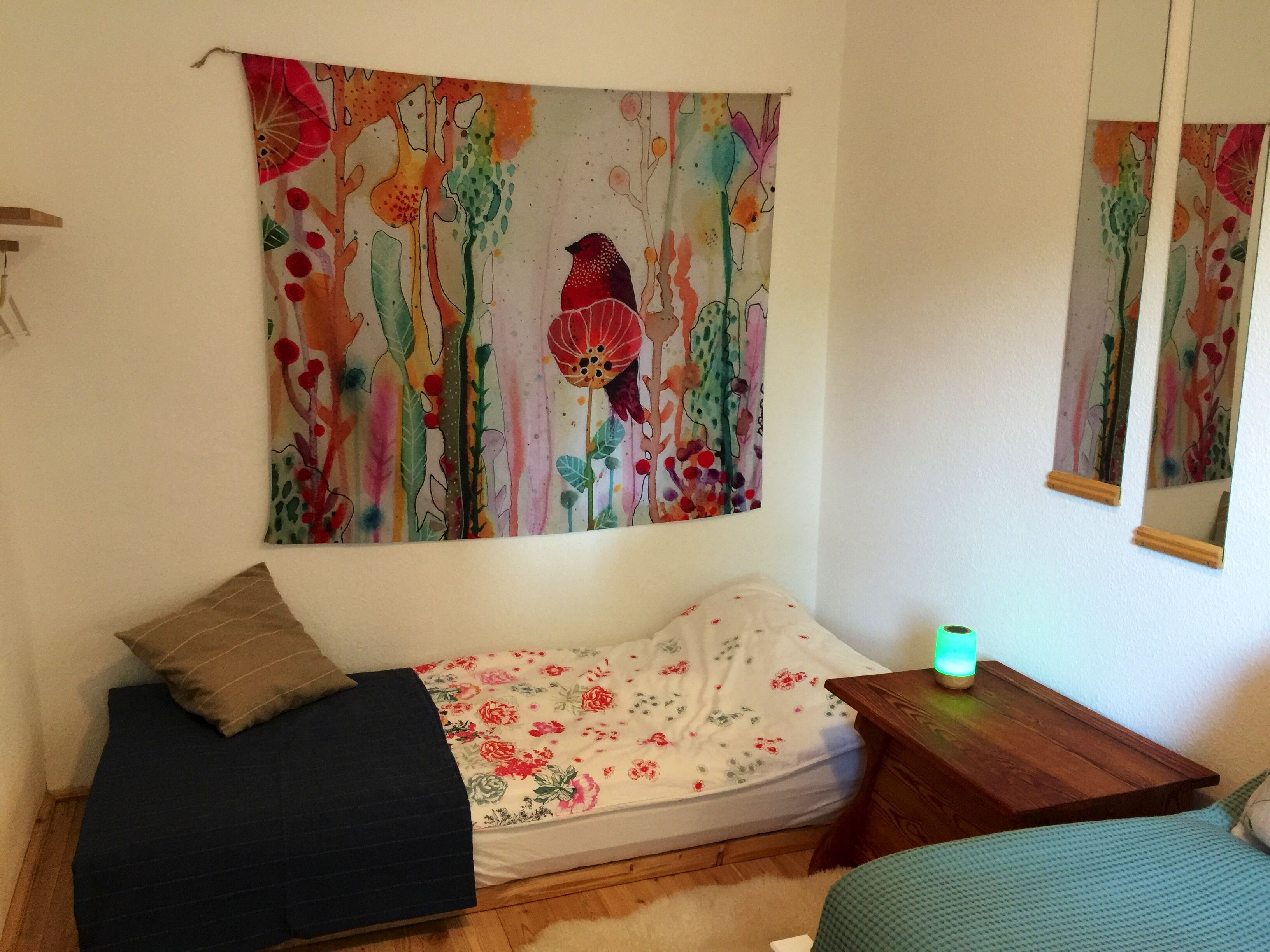 Ein kleines Zimmer mit einem Bett, einer Sitzbank, einem Tisch und einem farbenfrohen Wandteppich.
