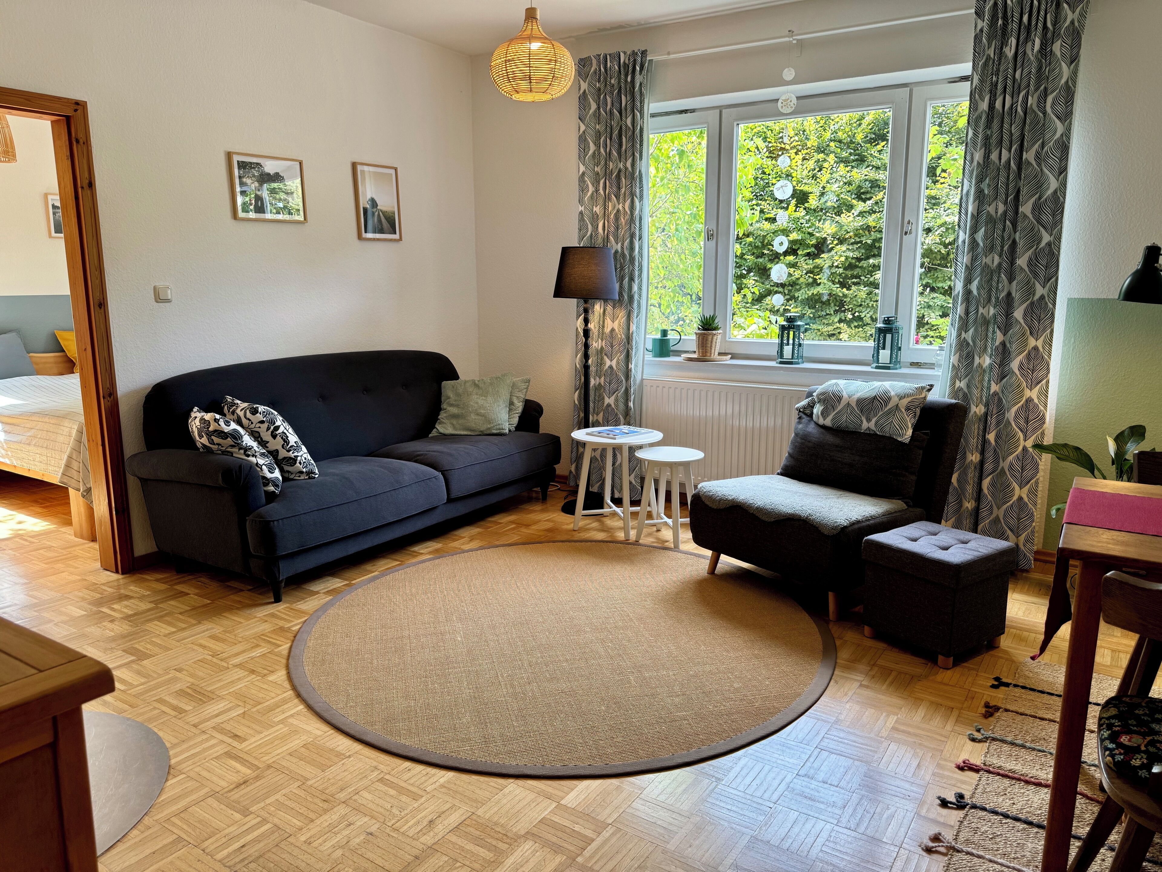 Een woonkamer met een donkere bank, een rond tapijt, een klein rond tafeltje en een raam met gordijnen.