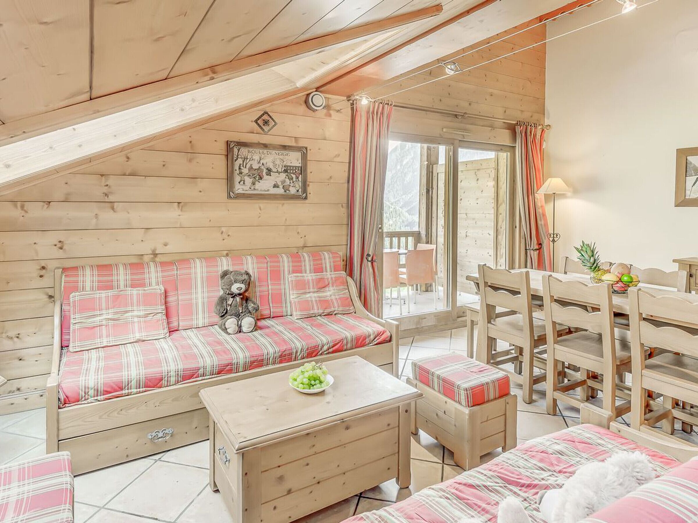 L’intérieur chaleureux d’un chalet avec un plancher en bois, un canapé à carreaux, une petite table sur laquelle se trouve un saladier rempli de fruits, et un coin repas avec des chaises et une table en bois.