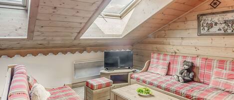 A cozy room with a sloped ceiling, wooden walls, a television, a sofa, a small table, and a window.