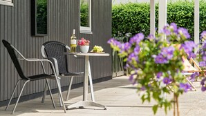 Eine Terrasse im Freien mit zwei Stühlen, einem Tisch mit einer Flasche und Blumen sowie Blick auf einen Heckenstreifen.