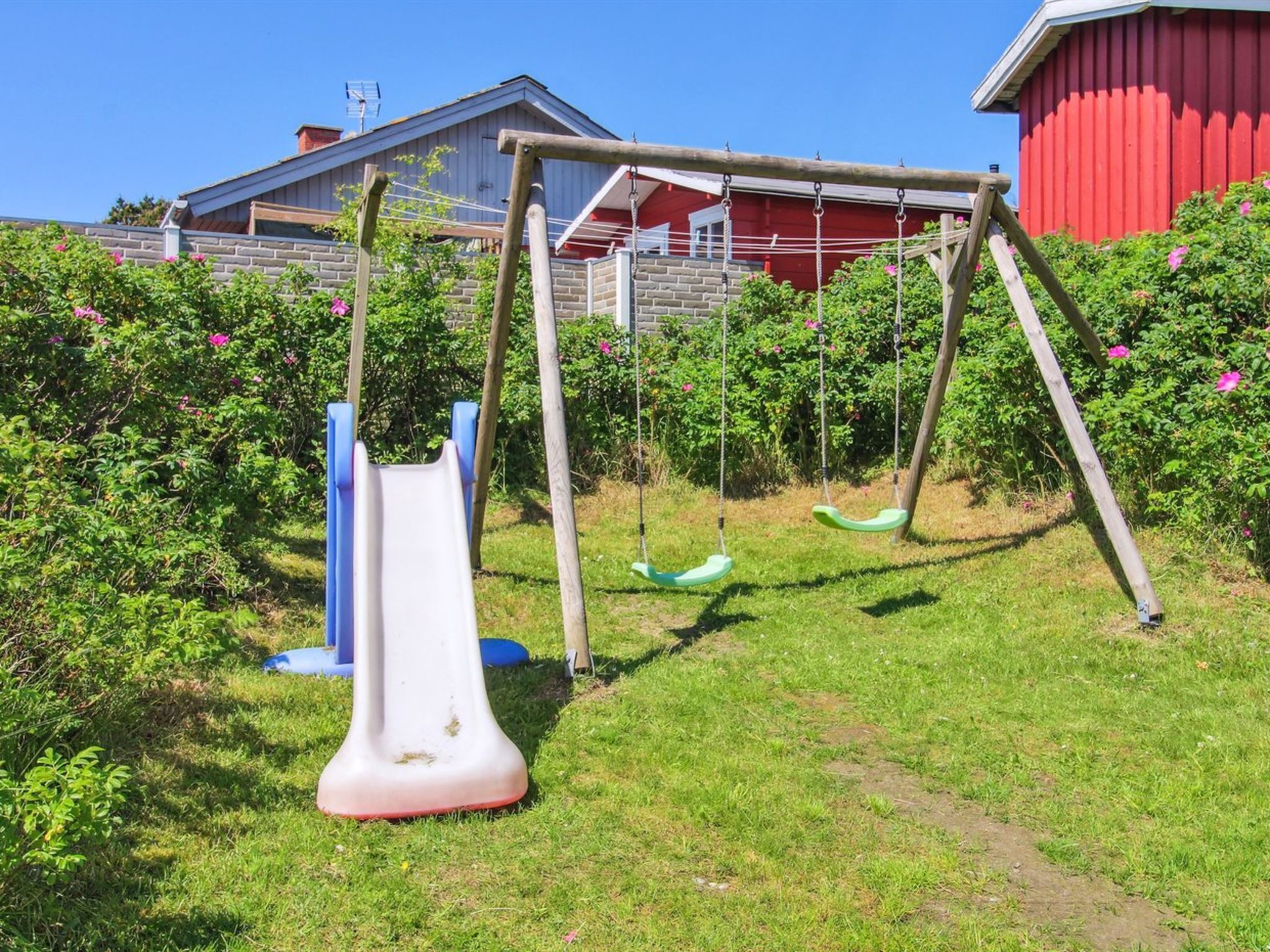 Un parc infantil amb tobogan i cadira de balanceig, envoltat de vegetació i un edifici vermell al fons.