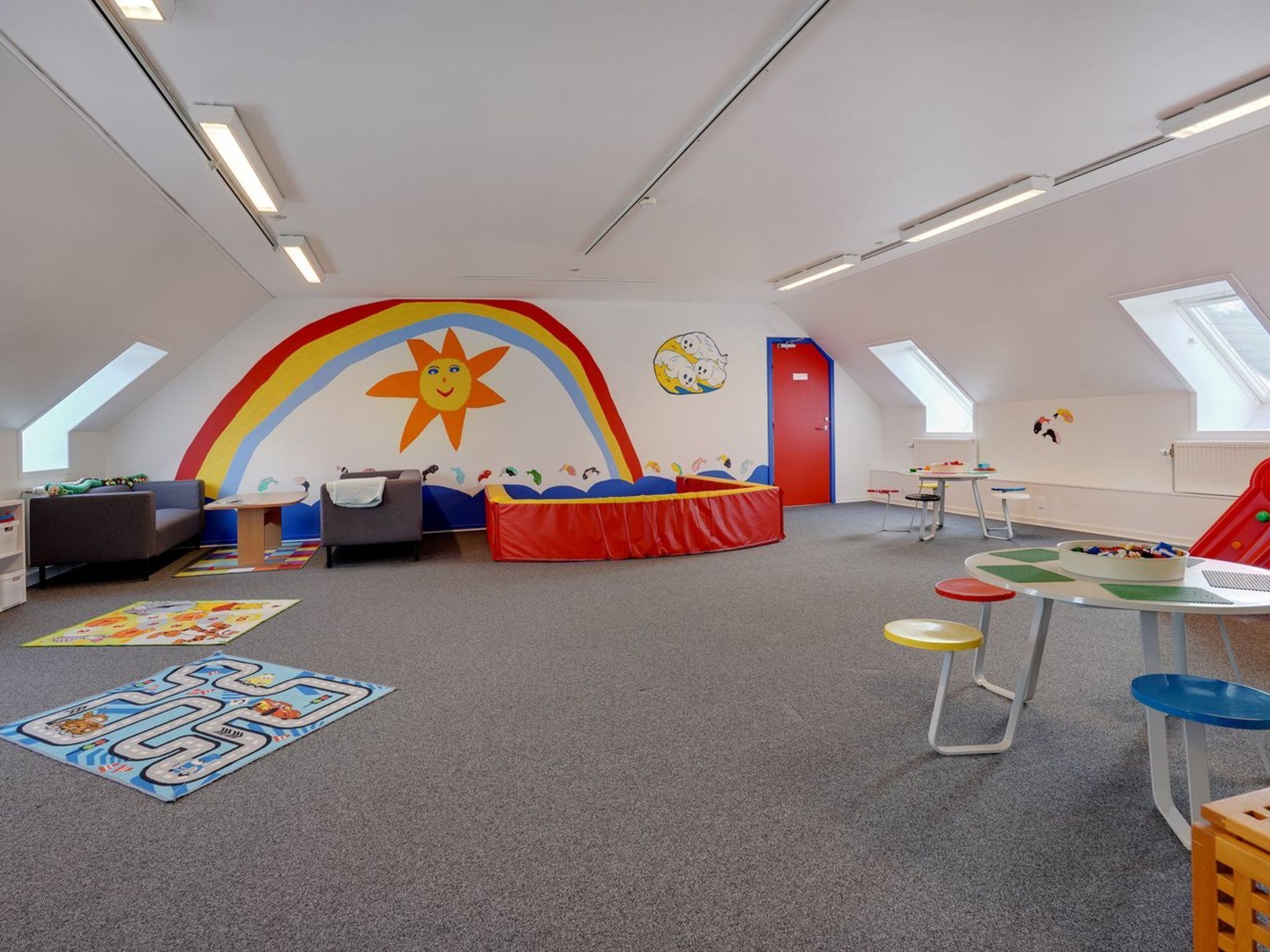 Een kinderspeelkamer met een kleurrijke muurschildering, een box, een tafel vol speelgoed en een bank.
