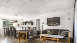 A spacious living room with a wooden floor, a black leather sofa, a dining area with chairs, and a flat-screen TV mounted on the wall.