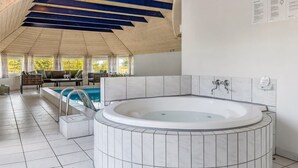 A modern indoor swimming pool area with a hot tub, wooden ceiling beams, and a seating area with a view of trees.