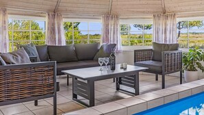 A covered outdoor seating area with a pool, featuring a sofa, chairs, and a table with glasses and a bottle.
