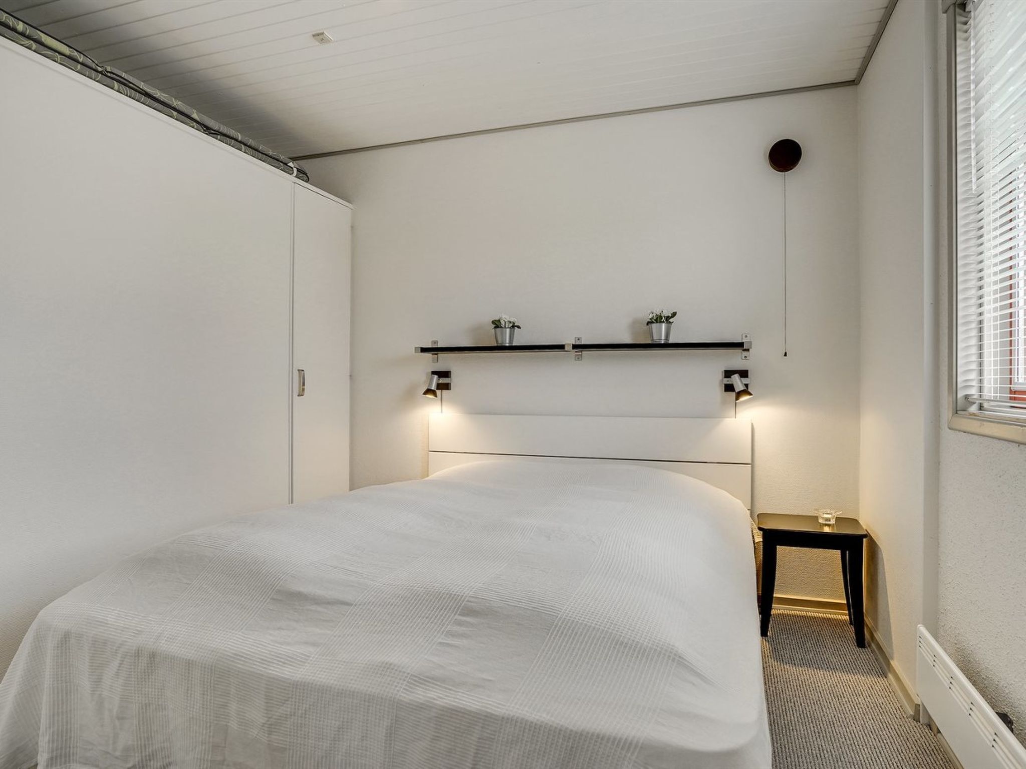 Une chambre à coucher avec un lit, des tables de chevet, une étagère avec des plantes et un radiateur.