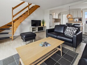 A modern living room with a wooden staircase, a black leather sofa, a wooden coffee table, and a kitchen in the background.