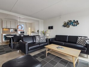 A modern living room with a black leather sofa, a wooden coffee table, and a black leather ottoman. There is a kitchen in the background with white cabinets and a hanging pendant light.