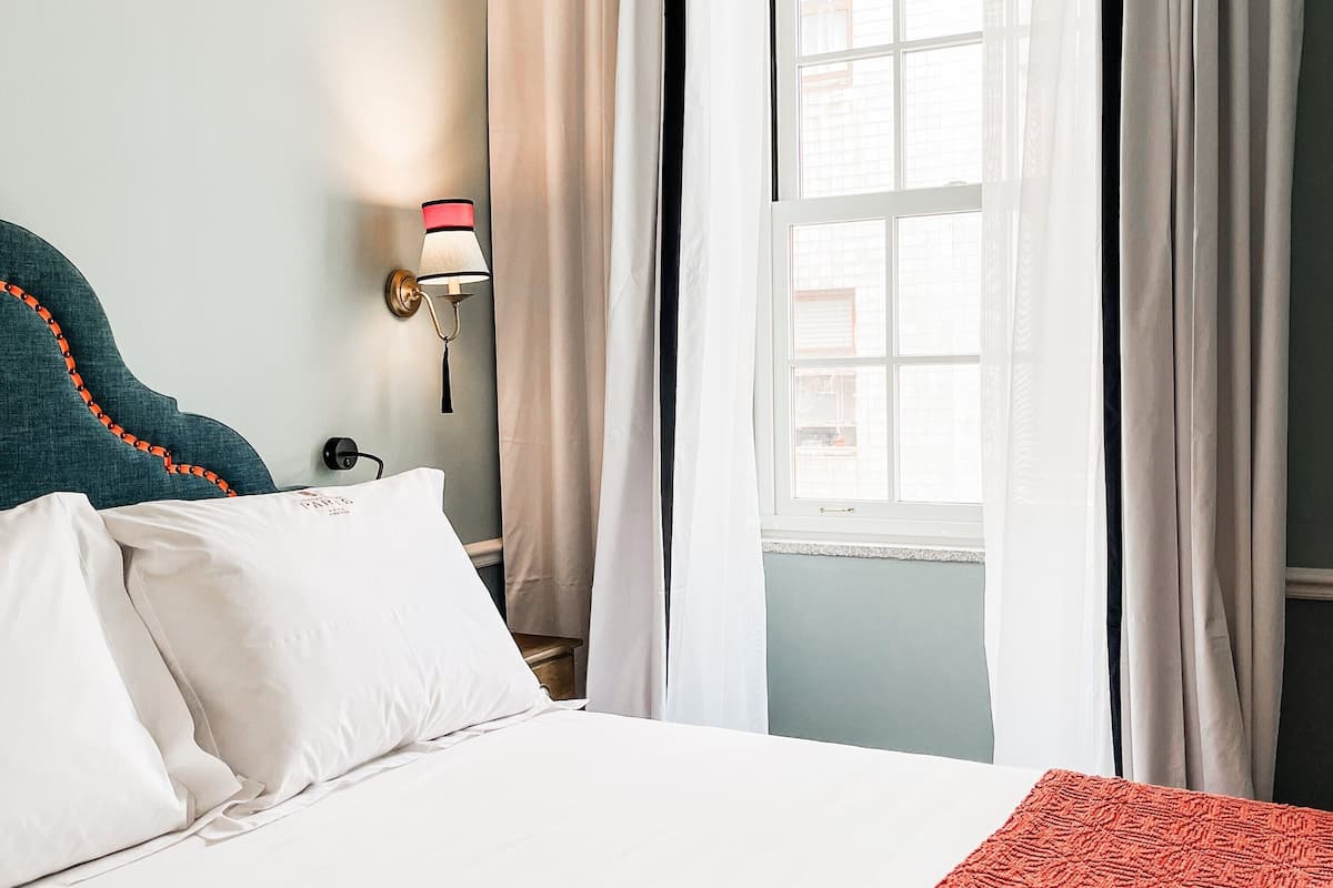 A neatly made bed with white and red bedding, a teal headboard, and a window with sheer curtains.